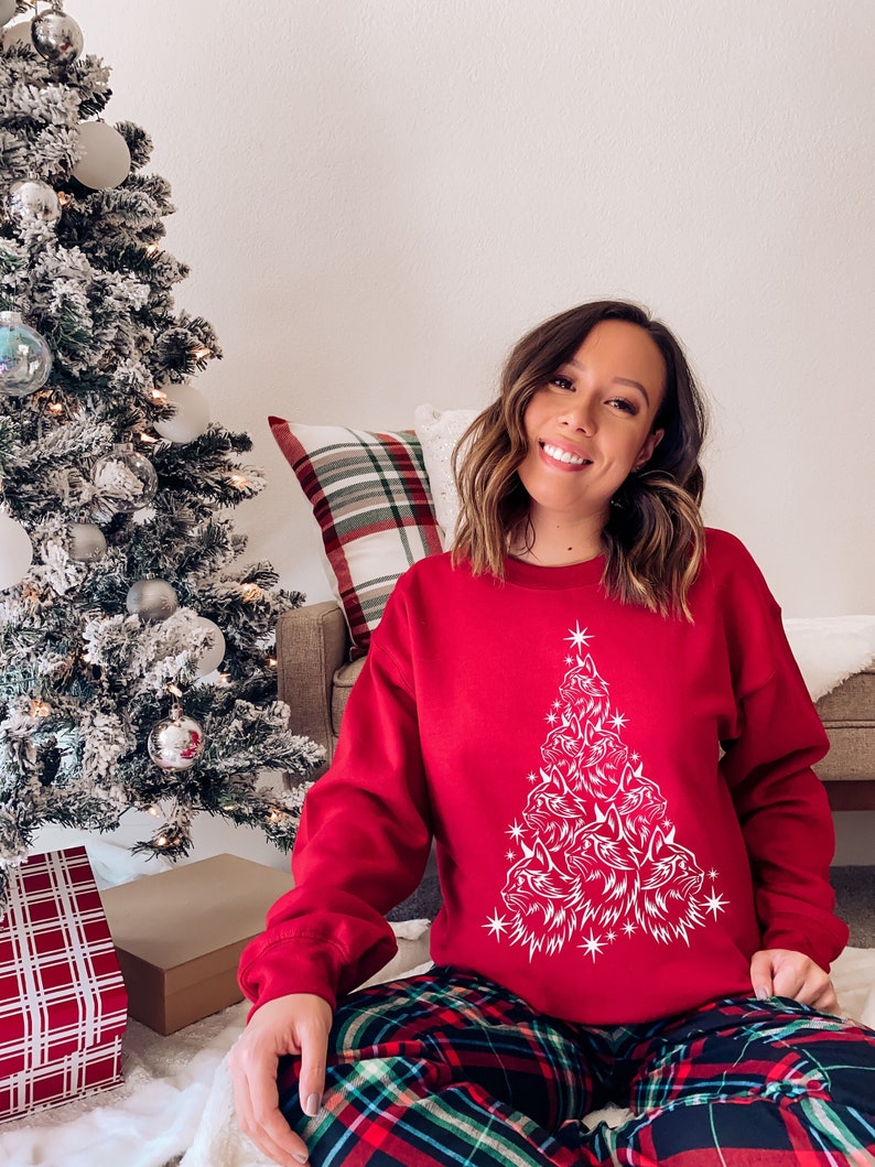 May include: A red sweatshirt featuring a white Christmas tree design made up of cat faces. The tree is decorated with white stars. The person wearing the sweatshirt is sitting on a plaid blanket.