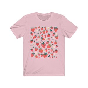 K&ouml;nnte beinhalten: Ein pinkfarbenes T-Shirt mit einem Muster aus roten Erdbeeren und wei&szlig;en Bl&uuml;ten. Die Erdbeeren sind in einem zuf&auml;lligen Muster angeordnet und haben unterschiedliche Gr&ouml;&szlig;en.