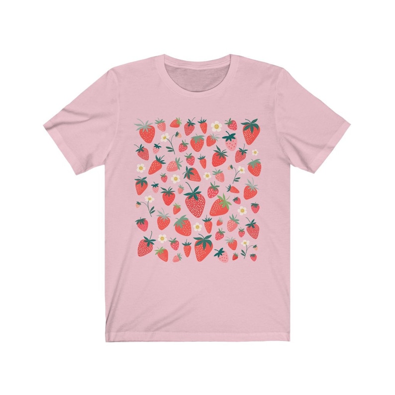 K&ouml;nnte beinhalten: Ein pinkfarbenes T-Shirt mit einem Muster aus roten Erdbeeren und wei&szlig;en Bl&uuml;ten. Die Erdbeeren sind in einem zuf&auml;lligen Muster angeordnet und haben unterschiedliche Gr&ouml;&szlig;en.