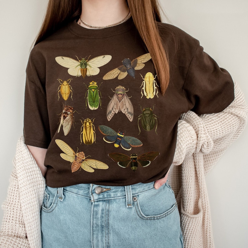 May include: A brown t-shirt featuring a vintage insect illustration print. The print depicts a variety of insects, including beetles, butterflies, and moths, in shades of green, brown, yellow, and black.