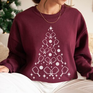 K&ouml;nnte beinhalten: Ein weinroter Pullover mit einem wei&szlig;en Design eines Weihnachtsbaums aus Pickleball-Schl&auml;gern und B&auml;llen.