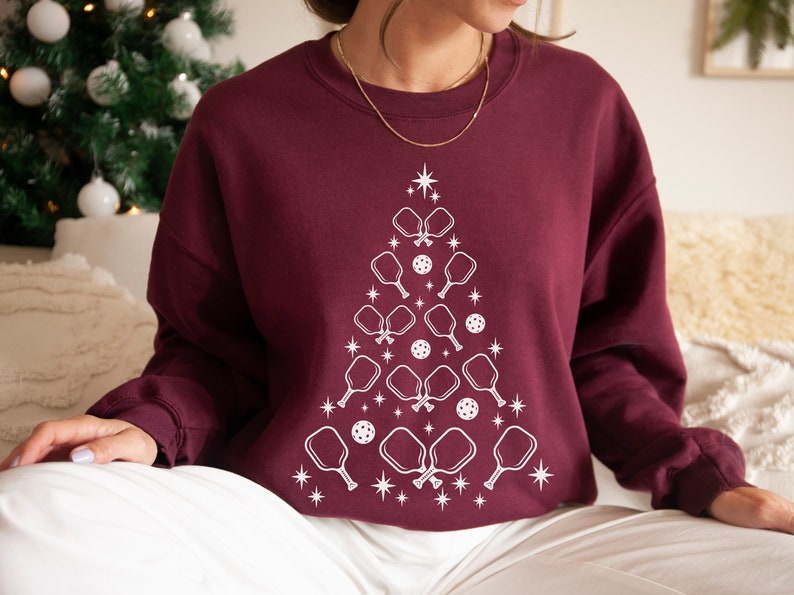 K&ouml;nnte beinhalten: Ein weinroter Pullover mit einem wei&szlig;en Design eines Weihnachtsbaums aus Pickleball-Schl&auml;gern und B&auml;llen.
