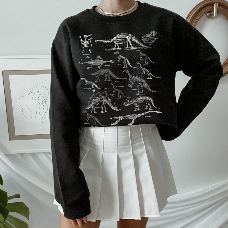 May include: Black cropped sweatshirt with a white print of dinosaur skeletons. The sweatshirt has a crew neck and long sleeves.