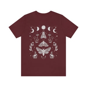 Puede incluir: Una camiseta color granate con un dise&ntilde;o gr&aacute;fico blanco que presenta polillas, setas y lunas. El dise&ntilde;o est&aacute; centrado en la camiseta y tiene un patr&oacute;n sim&eacute;trico.