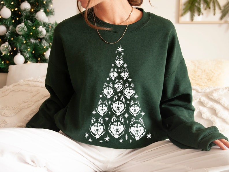 Puede incluir: Una sudadera verde con un dise&ntilde;o de &aacute;rbol de Navidad blanco con m&uacute;ltiples caras de perros husky. El &aacute;rbol est&aacute; decorado con estrellas blancas.