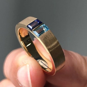 Puede incluir: Un anillo de banda de oro con tres piedras preciosas rectangulares. Las piedras preciosas son azules, azul claro y azul oscuro. El anillo está grabado con el texto "DiamondCreationArt".