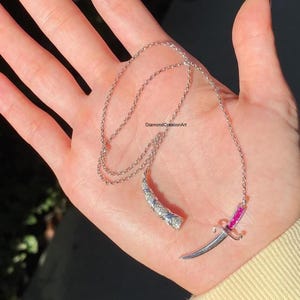 Könnte beinhalten: Eine silberne Halskette mit zwei Anhängern. Ein Anhänger ist eine gebogene Hornform mit kleinen weißen Steinen und blauen Akzenten. Der andere Anhänger ist ein kleiner Dolch mit einem rosa Edelstein am Griff. Die Halskette wird auf einer Hand präsentiert.