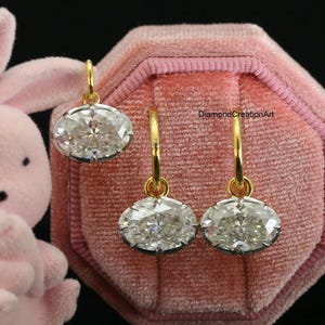 May include: A pair of oval-cut clear gemstone earrings with gold-toned hoops, displayed on a pink velvet octagonal jewellery box next to a small pink rabbit figurine. The text DiamondCreationArt is visible on the velvet surface of the box.
