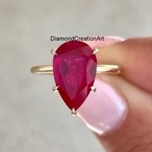 May include: A pear-shaped ruby gemstone set in a gold ring. The ring is on a finger.