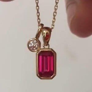May include: A gold necklace featuring a rectangular emerald-cut red gemstone pendant and a smaller round white gemstone charm. The pendant and charm are suspended from a delicate gold chain. The text DiamondCreationArt is visible in the image.