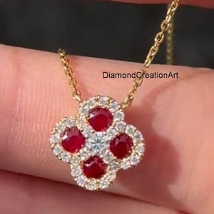 May include: A gold necklace with a four-leaf clover pendant. The pendant features four red gemstones encircled by a halo of small white gemstones. The necklace is held by a person.