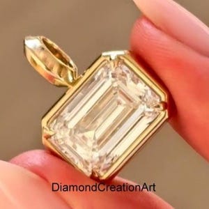 Pode incluir: Um pingente de ouro com uma grande pedra preciosa retangular e transparente. A pedra é engastada em uma moldura de ouro. O pingente é segurado entre dois dedos. O texto "DiamondCreationArt" é visível.