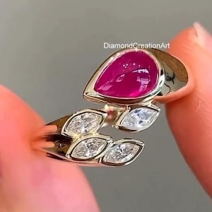 May include: A gold ring featuring a large pear-shaped deep pink gemstone and three smaller marquise-cut clear stones arranged in a leaf-like pattern. The band is open-ended, and the text DiamondCreationArt is visible above the ring.