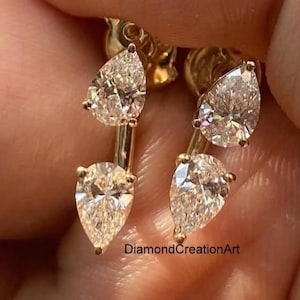 Op de afbeelding: Een paar gouden oorbellen, elk met twee verticaal geplaatste, peervormig geslepen heldere stenen. De stenen zijn gezet in een gouden metalen frame, wat zorgt voor een hangend ontwerp. De tekst DiamondCreationArt is in kleine letters zichtbaar.