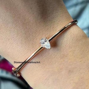 May include: A rose gold bangle bracelet with a single pear-shaped clear gemstone.