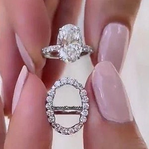 May include: A close-up of a silver engagement ring with a large oval-shaped diamond. The ring has a halo setting with smaller diamonds surrounding the center stone. The ring is held by a hand with manicured nails.