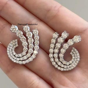 Puede incluir: Un par de pendientes plateados con tres filas curvas y superpuestas de piedras redondas, claras y facetadas. El diseño crea una forma de espiral en abanico. El texto DiamondCreationArt es visible en la esquina superior izquierda.