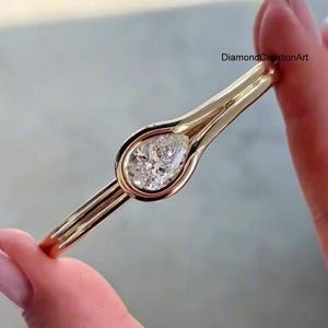 May include: A gold ring featuring a pear-shaped diamond set in a bezel setting. The band splits into two parallel lines that frame the central stone. The text DiamondCreationArt is visible in the upper right corner of the image.