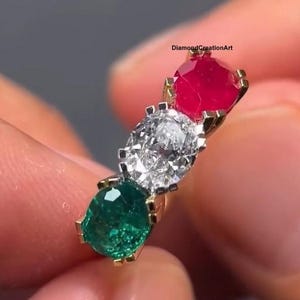 Könnte beinhalten: Ein goldener Ring mit drei Edelsteinen: einem roten Rubin, einem klaren Diamanten und einem grünen Smaragd. Die Edelsteine sind in einer Reihe angeordnet. Der Ring wird von einer Person gehalten. Der Text "DiamondCreationArt" ist sichtbar.