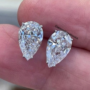 May include: A pair of pear-shaped diamond stud earrings. The earrings are set in a light-coloured metal and have a brilliant cut. The earrings are displayed on a light-coloured background. The text "DiamondCreationArt" is visible.