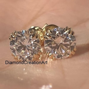 May include: A pair of round, clear diamond stud earrings with gold settings. The earrings are set in a prong setting and are displayed on a light pink background. The text "DiamondCreationArt" is visible.