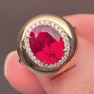May include: A gold ring featuring a large, oval-cut ruby gemstone. The ruby is surrounded by a halo of small, sparkling diamonds. The ring's band is a simple, polished gold.