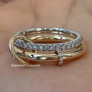 Op de afbeelding: Een ring met meerdere banden, bestaande uit een combinatie van gepolijst geelgoud en wit metaal. Eén band is bezet met kleine, heldere, rondgeslepen stenen. De banden zijn in elkaar verstrengeld in een cirkelvormig ontwerp. Tekst DiamondCreationArt is zichtbaar.