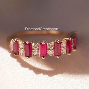 May include: A gold band ring featuring alternating rectangular red gemstones and small white gemstones. The ring is set against a soft, neutral background, highlighting the vibrant colors of the stones. The text "DiamondCreationArt" is visible.