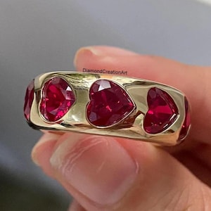 May include: A gold-toned band ring featuring a series of vibrant, deep red heart-shaped stones set flush into the metal surface. The ring displays the text DiamondCreationArt in small black lettering across the top center of the gold band.