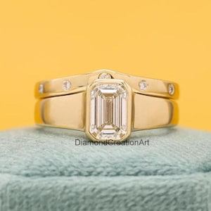 Puede incluir: Un anillo de compromiso de oro con un gran diamante central de talla esmeralda y diamantes más pequeños en la banda. El anillo está sobre un cojín de terciopelo azul.