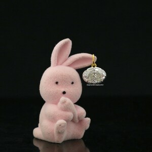 May include: A small, light pink, flocked bunny figurine sitting upright. The bunny has two black eyes and a small black nose. An oval-shaped, clear faceted gemstone pendant with a gold-coloured metal bail is suspended from one of the bunny's ears.