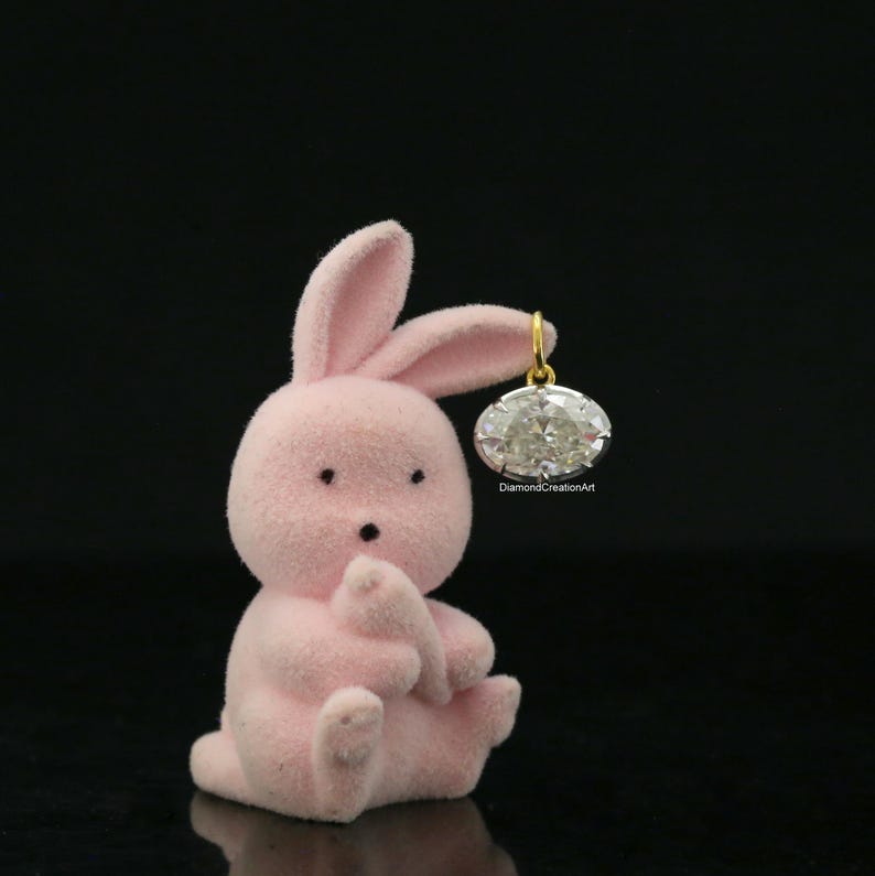 May include: A small, light pink, flocked bunny figurine sitting upright. The bunny has two black eyes and a small black nose. An oval-shaped, clear faceted gemstone pendant with a gold-coloured metal bail is suspended from one of the bunny's ears.