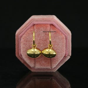 May include: A pair of gold hoop earrings featuring smooth, polished, oval-shaped charms that hang from thin circular hoops. The earrings are displayed against a pink velvet octagonal jewellery box with the text DiamondCreationArt visible.