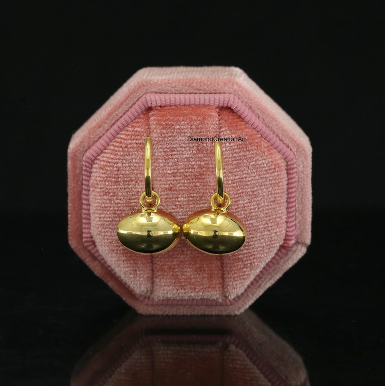 May include: A pair of gold hoop earrings featuring smooth, polished, oval-shaped charms that hang from thin circular hoops. The earrings are displayed against a pink velvet octagonal jewellery box with the text DiamondCreationArt visible.