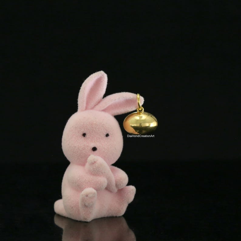 May include: A small, light pink flocked bunny figurine sitting upright. The bunny has two long ears, one of which is bent, and a small, shiny gold-coloured metal bell charm attached to it. The text DiamondCreationArt is printed in small black letters.