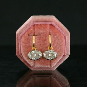 May include: A pair of drop earrings featuring oval-cut clear stones set in gold-toned metal hoops. The earrings are displayed in a pink velvet octagonal jewellery box. The text DiamondCreationArt is visible in the centre of the box.