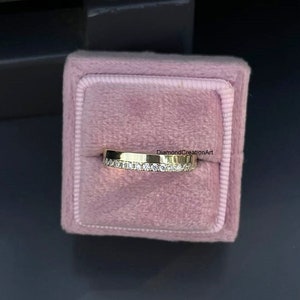 K&ouml;nnte beinhalten: Ein goldener Bandring mit einer Reihe kleiner, klarer Edelsteine, die in die obere H&auml;lfte eingesetzt sind. Der Ring wird in einer rosa Samt-Ringbox pr&auml;sentiert. Der Text "DiamondCreationArt" ist sichtbar.