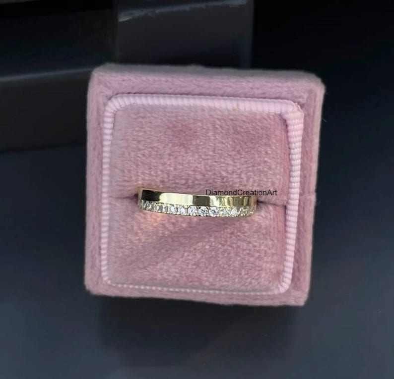 K&ouml;nnte beinhalten: Ein goldener Bandring mit einer Reihe kleiner, klarer Edelsteine, die in die obere H&auml;lfte eingesetzt sind. Der Ring wird in einer rosa Samt-Ringbox pr&auml;sentiert. Der Text "DiamondCreationArt" ist sichtbar.