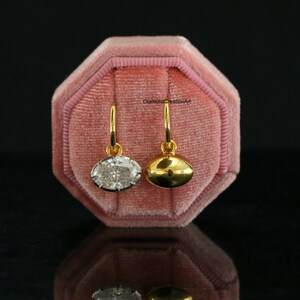 May include: Two gold hoop earrings with dangling charms. One charm is an oval-cut clear gemstone, and the other is a smooth, polished gold oval. The text DiamondCreationArt is visible. The earrings are displayed on a pink velvet octagonal box.