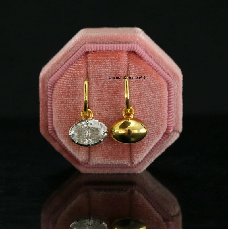 May include: Two gold hoop earrings with dangling charms. One charm is an oval-cut clear gemstone, and the other is a smooth, polished gold oval. The text DiamondCreationArt is visible. The earrings are displayed on a pink velvet octagonal box.