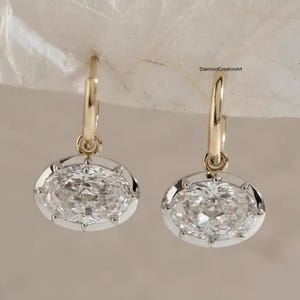 Op de afbeelding: Een paar gouden oorringen met ovale, heldere gefacetteerde stenen in een klauwzetting. De oorbellen hangen aan dunne gouden ringen. De tekst DiamondCreationArt is zichtbaar in de rechterbovenhoek van de afbeelding.