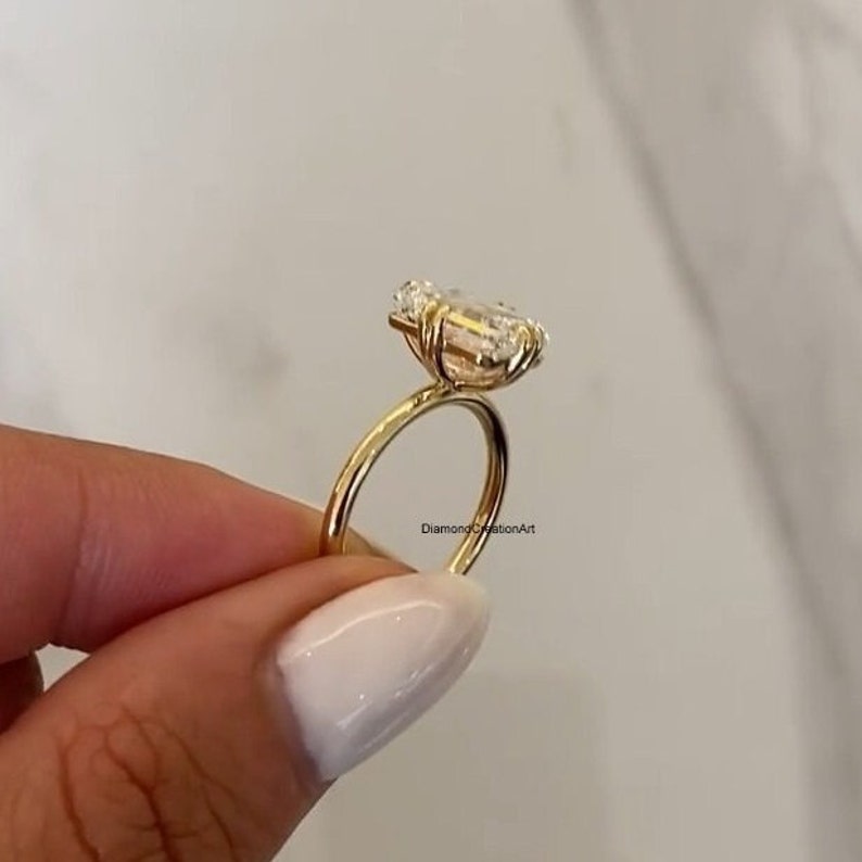 May include: A gold ring featuring a large, clear emerald-cut diamond. The ring is set on a thin band and the diamond is held in place by four prongs. The ring is being held by a hand.