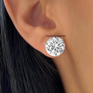 May include: A close-up of a sparkling, round-cut diamond stud earring. The earring is set in a silver or white gold setting and is worn on an ear. The image is well-lit, highlighting the brilliance of the diamond. The text "DiamondCreationArt" is visible.