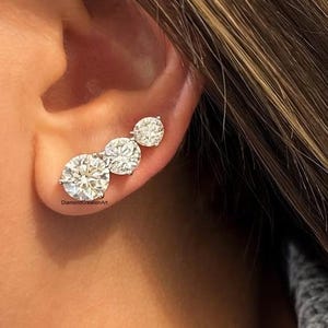 May include: A close-up of a three-stone diamond earring set. The earring features three round, brilliant-cut diamonds of varying sizes, set in a silver or white gold setting. The diamonds sparkle against the skin.