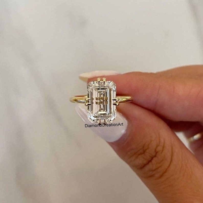 May include: A gold ring featuring a large, clear emerald-cut diamond. The diamond is set in a delicate, prong setting. The ring is on a finger.