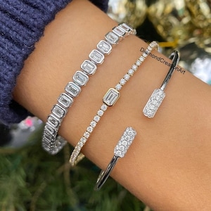 Puede incluir: Tres pulseras de diamantes. Una es una pulsera plateada con diamantes rectangulares. La segunda es una pulsera dorada con pequeños diamantes redondos y un diamante rectangular. La tercera es una pulsera de puño plateada con detalles de diamantes.