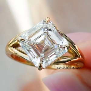 Puede incluir: Primer plano de un anillo de oro con una gran piedra preciosa transparente de talla cuadrada. El anillo tiene un diseño de caña dividida. La piedra preciosa está facetada, reflejando la luz. El anillo está en la mano de una persona.