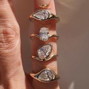 Puede incluir: Cuatro anillos de oro con piedras preciosas claras se muestran en un dedo. Las piedras preciosas tienen forma de pera y ovalada. Los anillos tienen un diseño simple y elegante. Los anillos son probablemente para compromisos u ocasiones especiales.