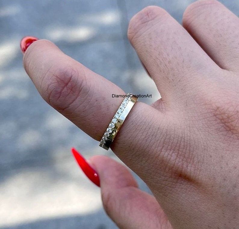 K&ouml;nnte beinhalten: Ein goldener Bandring mit einer Reihe kleiner, klarer Edelsteine an einer Kante. Der Ring wird an einem Finger getragen, mit dem Text "DiamondCreationArt" im Hintergrund.