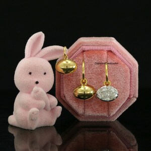 May include: A pink flocked bunny figurine stands next to a pink velvet octagonal jewellery box containing three gold hoop earrings. Two earrings feature smooth gold oval charms, while the third earring displays a faceted clear oval stone.